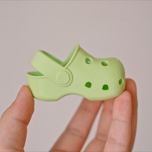 May include: A light green plastic clog with a strap and holes on the top. The clog is being held in a hand.