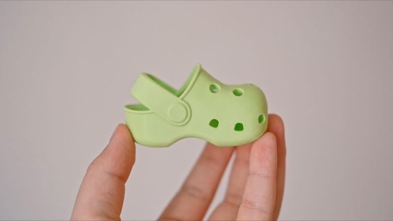 May include: A light green plastic clog with a strap and holes on the top. The clog is being held in a hand.