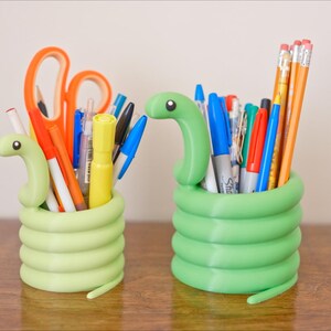 May include: Two green snake-shaped desk organizers filled with pens, pencils, and scissors. The organizers are made of a light green material and have a coiled body design. The desk organizers are on a brown wooden surface.