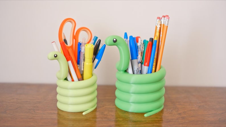 May include: Two green snake-shaped desk organizers filled with pens, pencils, and scissors. The organizers are made of a light green material and have a coiled body design. The desk organizers are on a brown wooden surface.