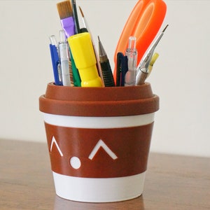 May include: A brown and white ceramic cup with a cartoon face and a white rim, filled with various writing and art supplies, including pens, pencils, paintbrushes, and a pair of scissors.