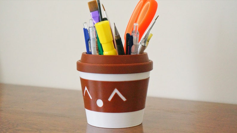 May include: A brown and white ceramic cup with a cartoon face and a white rim, filled with various writing and art supplies, including pens, pencils, paintbrushes, and a pair of scissors.