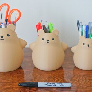 May include: Three brown bear-shaped ceramic pencil holders, each with a different number of pens and pencils inside. The largest holder has scissors and paintbrushes, the middle holder has a variety of pens, and the smallest holder has a bunch of pens and a green highlighter.