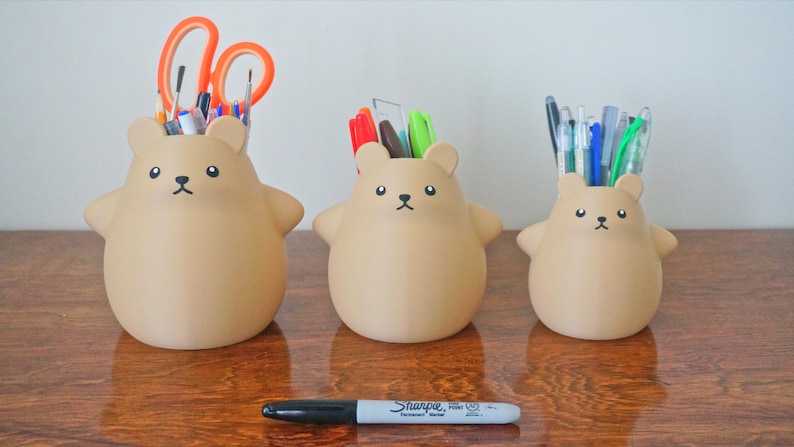 May include: Three brown bear-shaped ceramic pencil holders, each with a different number of pens and pencils inside. The largest holder has scissors and paintbrushes, the middle holder has a variety of pens, and the smallest holder has a bunch of pens and a green highlighter.