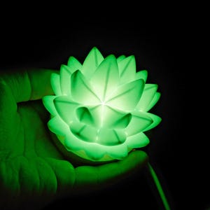 K&ouml;nnte beinhalten: Eine leuchtend gr&uuml;ne Lotusbl&uuml;ten-Lampe, die in einer Hand vor schwarzem Hintergrund gehalten wird. Die Lampe strahlt ein weiches, diffuses Licht aus, das das komplizierte Bl&uuml;tenblattdesign hervorhebt. Der Gesamteffekt ist ruhig und entspannend.
