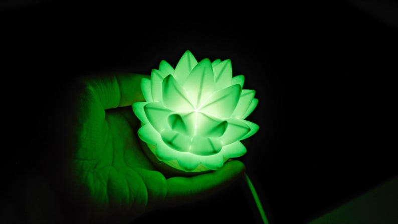 K&ouml;nnte beinhalten: Eine leuchtend gr&uuml;ne Lotusbl&uuml;ten-Lampe, die in einer Hand vor schwarzem Hintergrund gehalten wird. Die Lampe strahlt ein weiches, diffuses Licht aus, das das komplizierte Bl&uuml;tenblattdesign hervorhebt. Der Gesamteffekt ist ruhig und entspannend.
