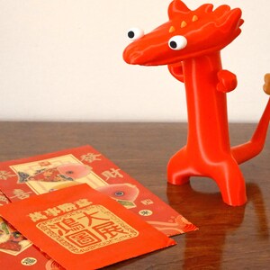 May include: A bright red, stylized dragon figurine with large white eyes and a small tail stands next to three red envelopes. The envelopes feature gold and red designs with Chinese characters, suggesting a cultural theme.