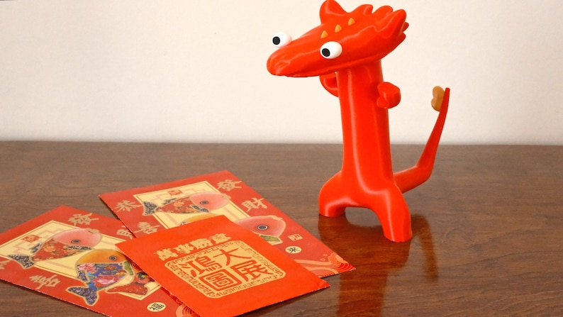 May include: A bright red, stylized dragon figurine with large white eyes and a small tail stands next to three red envelopes. The envelopes feature gold and red designs with Chinese characters, suggesting a cultural theme.