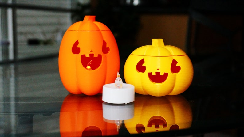 May include: Two Halloween jack-o'-lantern candle holders, one orange and one yellow, with a white tea light candle in between.