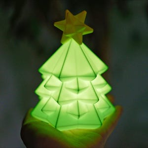 May include: A glowing green Christmas tree-shaped lamp with a yellow star on top. The lamp has a geometric design and is lit from within, creating a soft, ambient glow. The tree is held in a hand against a blurred background.