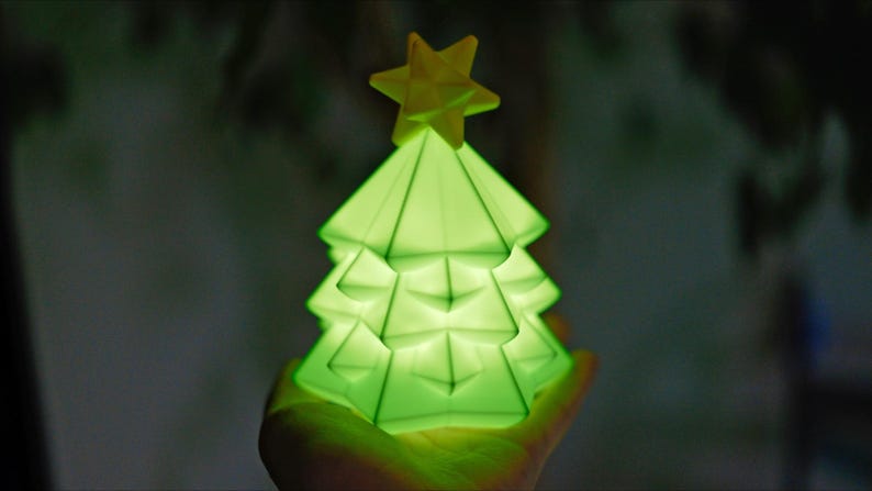 May include: A glowing green Christmas tree-shaped lamp with a yellow star on top. The lamp has a geometric design and is lit from within, creating a soft, ambient glow. The tree is held in a hand against a blurred background.