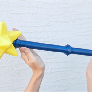 May include: A blue and yellow star-shaped wand. The wand has a bright yellow star at the top and a blue handle. The star is multi-faceted, and the handle has decorative rings. The wand is held in two hands.