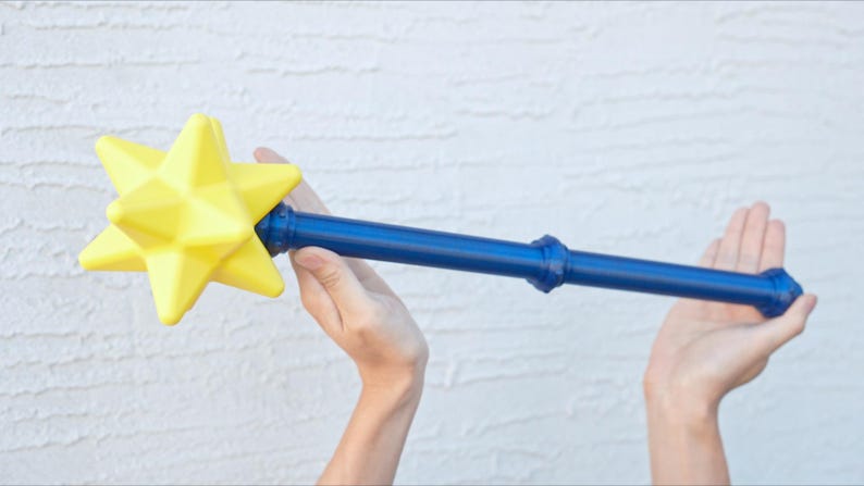 May include: A blue and yellow star-shaped wand. The wand has a bright yellow star at the top and a blue handle. The star is multi-faceted, and the handle has decorative rings. The wand is held in two hands.