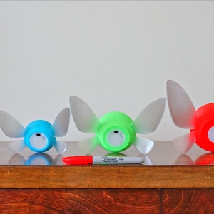 May include: Four different sized fairy lights, each with a different color: pink, blue, green, and red. The lights are shaped like a fairy with wings and a round body. The lights are on a wooden surface.