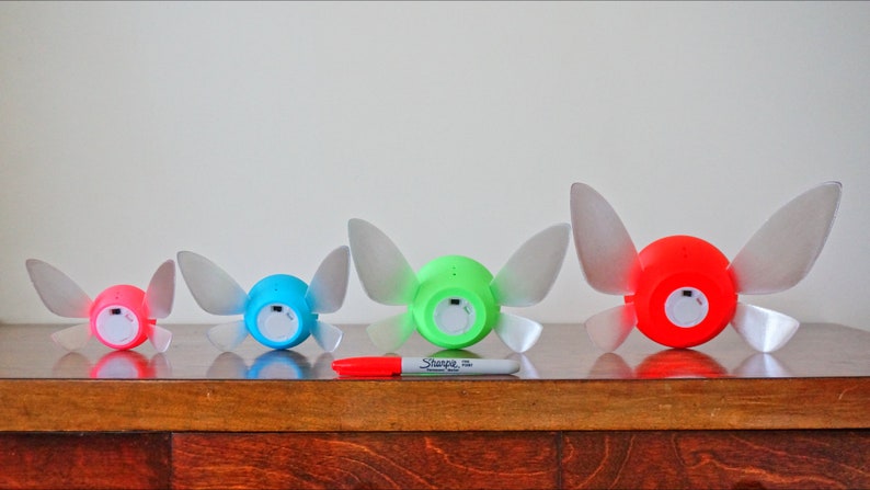 May include: Four different sized fairy lights, each with a different color: pink, blue, green, and red. The lights are shaped like a fairy with wings and a round body. The lights are on a wooden surface.