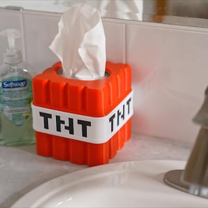 K&ouml;nnte beinhalten: Eine rote und wei&szlig;e Tissuebox in Form eines TNT-Blocks mit dem Text "THT" auf jeder Seite. Die Tissuebox steht auf einer wei&szlig;en Badezimmertheke.