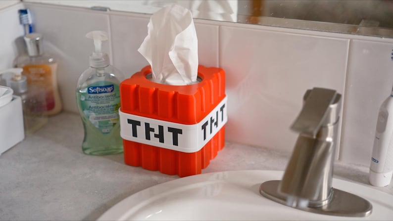 K&ouml;nnte beinhalten: Eine rote und wei&szlig;e Tissuebox in Form eines TNT-Blocks mit dem Text "THT" auf jeder Seite. Die Tissuebox steht auf einer wei&szlig;en Badezimmertheke.