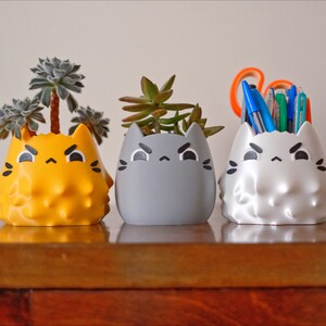 May include: Three ceramic cat-shaped planters, each with a different color and design. The planter on the left is yellow and has a succulent plant growing in it. The middle planter is gray and also has a succulent plant growing in it. The planter on the right is white and has a bunch of pens and pencils in it.