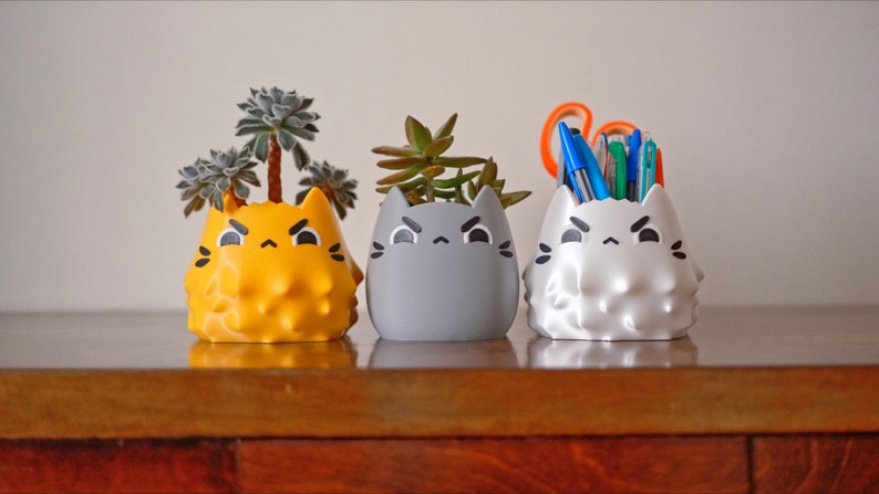 May include: Three ceramic cat-shaped planters, each with a different color and design. The planter on the left is yellow and has a succulent plant growing in it. The middle planter is gray and also has a succulent plant growing in it. The planter on the right is white and has a bunch of pens and pencils in it.