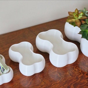 May include: Four white ceramic planters in different sizes, each with a unique, rounded, double-bubble shape. The two largest planters are filled with green succulents.