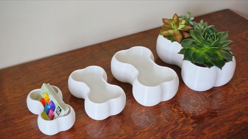 May include: Four white ceramic planters in different sizes, each with a unique, rounded, double-bubble shape. The two largest planters are filled with green succulents.