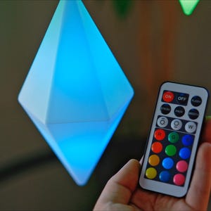 May include: A remote control with a variety of colored buttons, including red, green, blue, and yellow. The remote control is being held in a hand, and there are several diamond-shaped lights in the background.