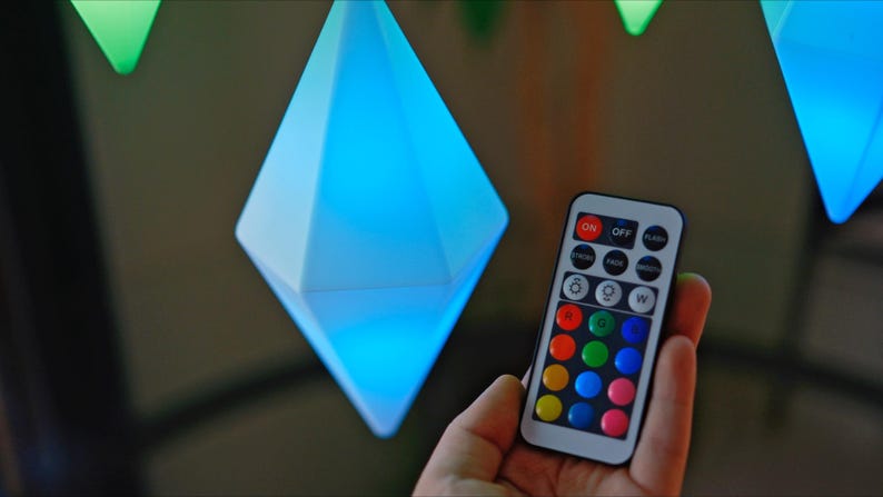 May include: A remote control with a variety of colored buttons, including red, green, blue, and yellow. The remote control is being held in a hand, and there are several diamond-shaped lights in the background.