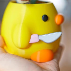 May include: A yellow ceramic container shaped like a cartoon duck, with a black eye, orange accents, and a knife. The container is held in a hand.