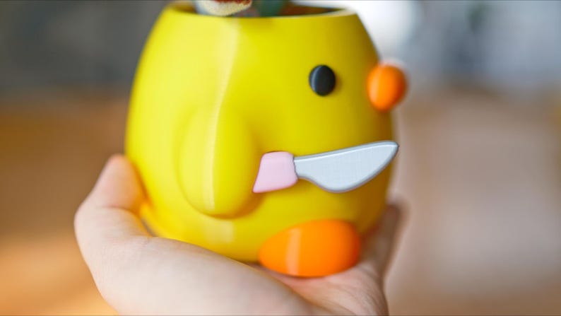May include: A yellow ceramic container shaped like a cartoon duck, with a black eye, orange accents, and a knife. The container is held in a hand.