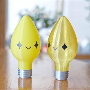 May include: Two decorative light bulbs with a whimsical design. One is bright yellow, the other is gold. Each bulb has a smiling face with diamond-shaped eyes and a curved mouth. The bulbs have silver bases and are set on a wooden table.