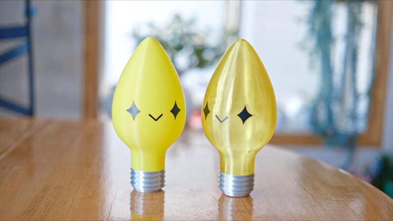 May include: Two decorative light bulbs with a whimsical design. One is bright yellow, the other is gold. Each bulb has a smiling face with diamond-shaped eyes and a curved mouth. The bulbs have silver bases and are set on a wooden table.