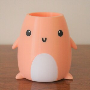 May include: A peach-coloured ceramic pencil holder shaped like a penguin with a white belly and black eyes and a smile.