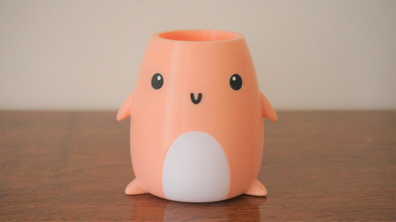 May include: A peach-coloured ceramic pencil holder shaped like a penguin with a white belly and black eyes and a smile.