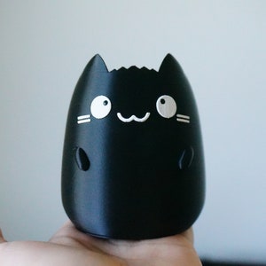 May include: A black, cat-shaped decorative item with white facial features, including eyes, whiskers, and a smiling mouth. The item has a rounded body and small, rounded ears. The item is held in a hand.