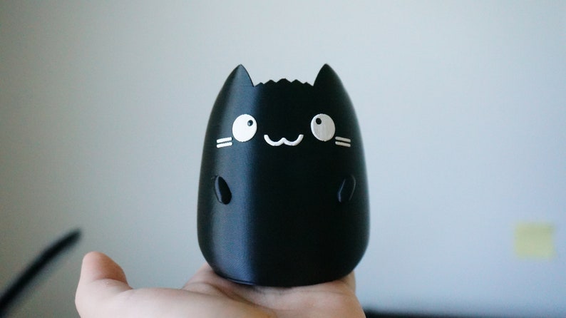 May include: A black, cat-shaped decorative item with white facial features, including eyes, whiskers, and a smiling mouth. The item has a rounded body and small, rounded ears. The item is held in a hand.
