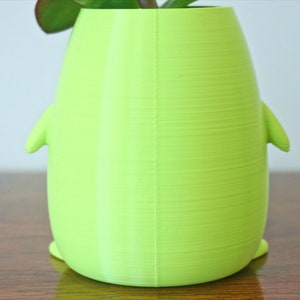 May include: A bright green, 3D printed planter shaped like a penguin. The planter has a smooth, textured surface and is sitting on a wooden table.