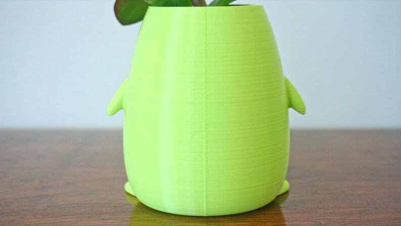 May include: A bright green, 3D printed planter shaped like a penguin. The planter has a smooth, textured surface and is sitting on a wooden table.