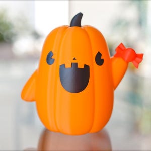 May include: A friendly orange pumpkin figurine with a black smiling face and a red bow. The pumpkin is holding out its right arm in a waving gesture.