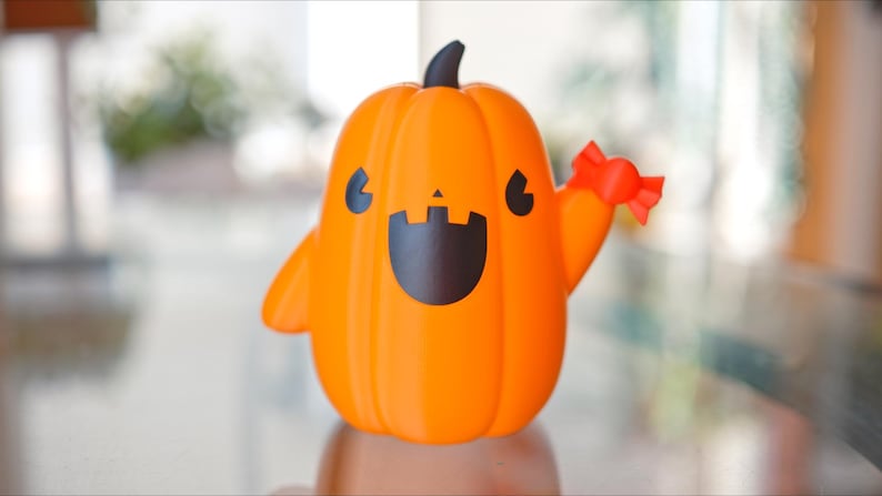 May include: A friendly orange pumpkin figurine with a black smiling face and a red bow. The pumpkin is holding out its right arm in a waving gesture.