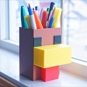 K&ouml;nnte beinhalten: Ein brauner, gelber und roter Minecraft-Stiftbeh&auml;lter in Form eines Huhns. Der Beh&auml;lter hat einen quadratischen Boden und ist mit verschiedenen Stiften und Bleistiften gef&uuml;llt.