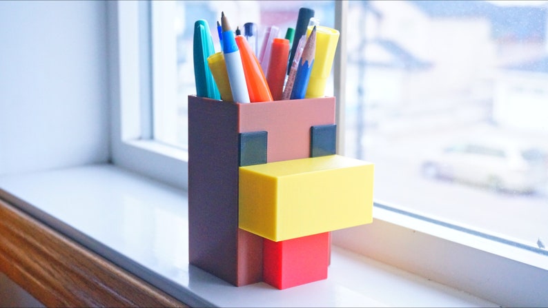 K&ouml;nnte beinhalten: Ein brauner, gelber und roter Minecraft-Stiftbeh&auml;lter in Form eines Huhns. Der Beh&auml;lter hat einen quadratischen Boden und ist mit verschiedenen Stiften und Bleistiften gef&uuml;llt.