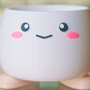 May include: White ceramic planter with a cute cartoon face. The planter has pink cheeks, black eyes, and a small black smile. The planter has two small feet.