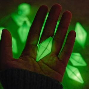 May include: A string of glowing green diamond-shaped lights. One of the lights is held in a hand, with other lights scattered in the background. The lights emit a soft, ambient glow, creating a unique and eye-catching display.