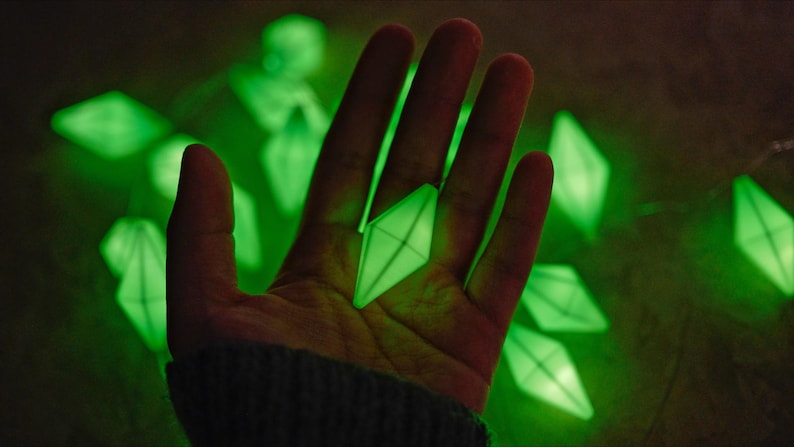 May include: A string of glowing green diamond-shaped lights. One of the lights is held in a hand, with other lights scattered in the background. The lights emit a soft, ambient glow, creating a unique and eye-catching display.