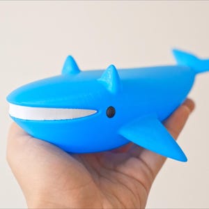May include: A bright blue whale figurine with a white mouth and black eyes. The whale has fins and a tail, and is held in a hand. The figurine is a toy or decorative item.