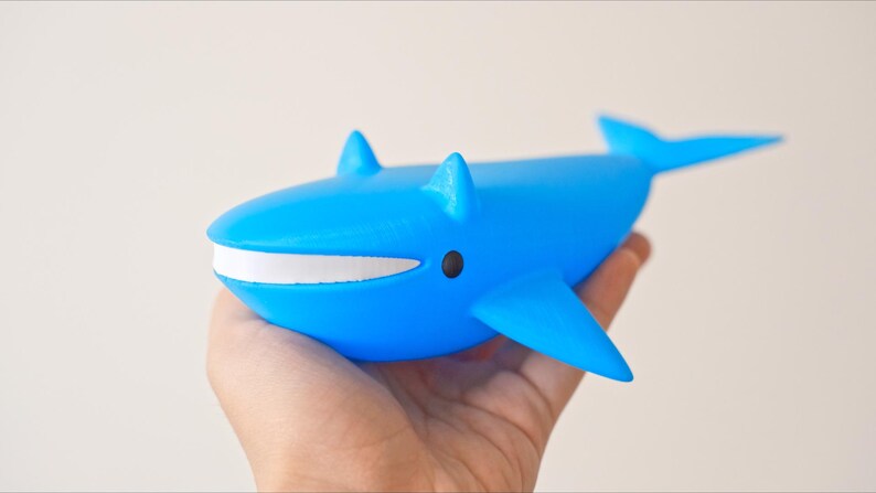 May include: A bright blue whale figurine with a white mouth and black eyes. The whale has fins and a tail, and is held in a hand. The figurine is a toy or decorative item.