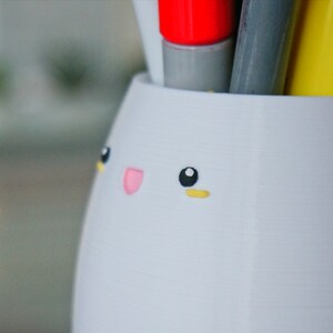 May include: White ceramic pencil holder shaped like a bunny with black eyes, a pink nose, and yellow eyebrows. The holder is filled with pens and markers.
