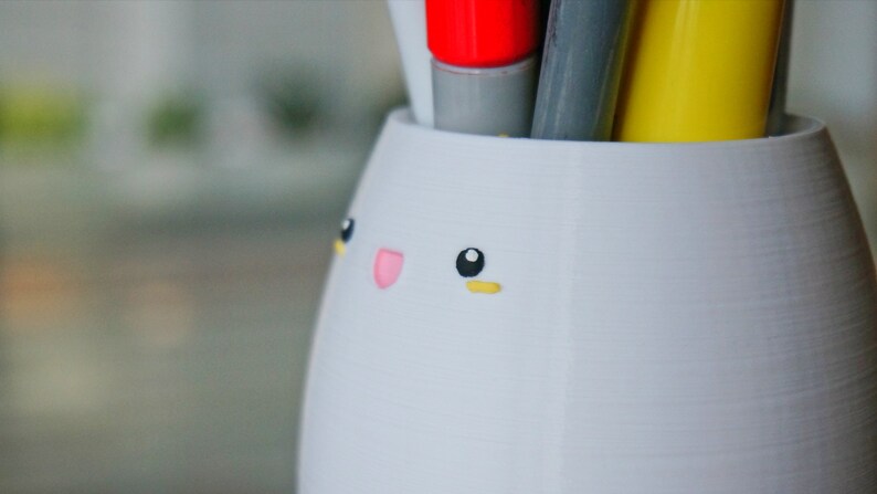 May include: White ceramic pencil holder shaped like a bunny with black eyes, a pink nose, and yellow eyebrows. The holder is filled with pens and markers.