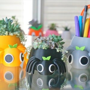 May include: A collection of whimsical spider-shaped planters in yellow, black, and gray, each holding succulents. The yellow and gray planters have a round body with eight legs and a face. The black planter has a similar design. The gray planter holds pens and pencils.
