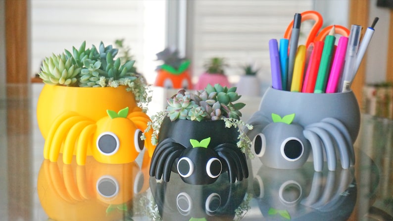 May include: A collection of whimsical spider-shaped planters in yellow, black, and gray, each holding succulents. The yellow and gray planters have a round body with eight legs and a face. The black planter has a similar design. The gray planter holds pens and pencils.